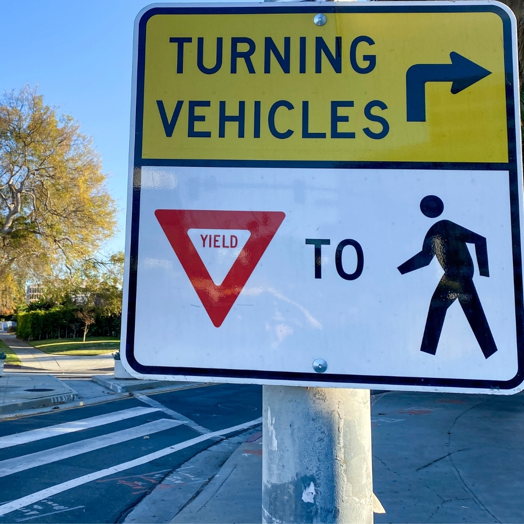 A traffic sign with yellow and black text reading "turning vehicles" and arrow pointing right to yield to pedestrians, a little walking man.