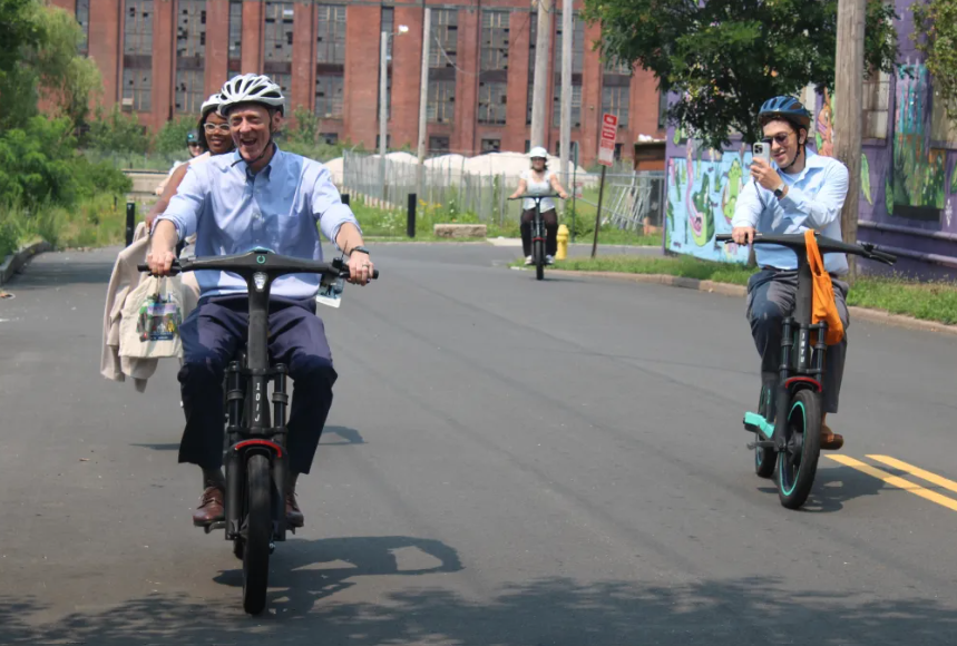 Two men and a woman wearing helmets ride seated scooters down a street in New Haven.