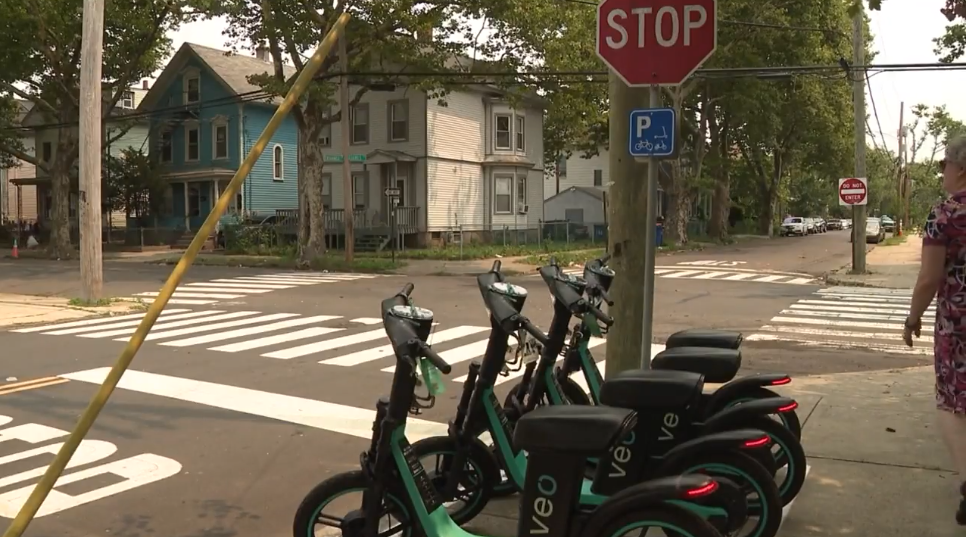 4 seated Veo scooters parked underneath a scooter and bike parking sign.